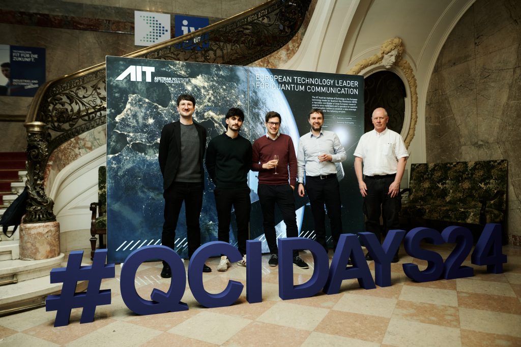 The core team of the KMS development. Picture taken at the QCI-Days 2024, which is shown in big letters. Historic staircase and AIT banner in the background.